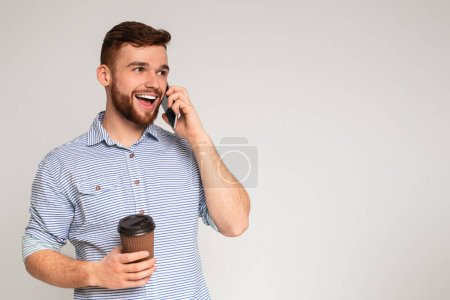 Photo for Handsome millennial guy talking on cellphone and holding coffee to go on studio background, panorama - Royalty Free Image