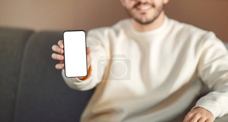 Téléchargez les photos : Coupé de l'homme est assis sur un canapé à la maison, tenant un téléphone portable avec écran blanc dans sa main, panorama - en image libre de droit