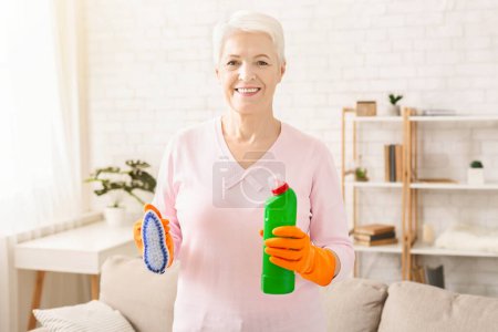 Senior woman is shown holding a bottle of cleaner and a rag in her hands. She appears to be ready to clean or wipe surfaces, displaying household chores in action.