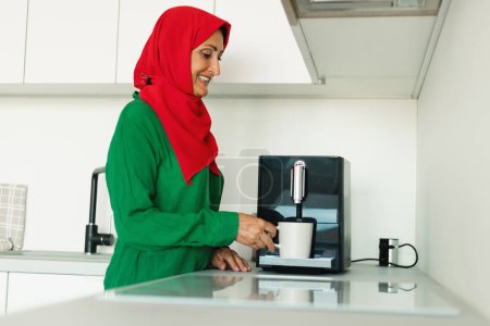 Téléchargez les photos : Une femme portant un hijab rouge et vert se tient dans une cuisine, versant de l'eau chaude dans une tasse à café. Elle semble concentrée alors qu'elle prépare sa boisson du matin. - en image libre de droit