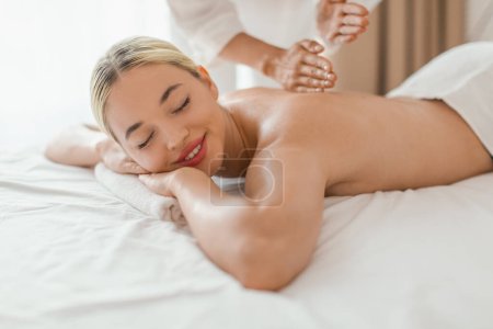 Eine zufriedene und friedliche Frau liegt mit dem Gesicht nach unten, während eine Masseurin eine entspannende Rückenmassage anbietet. Das Tageslicht taucht den Raum in ein sanftes Licht