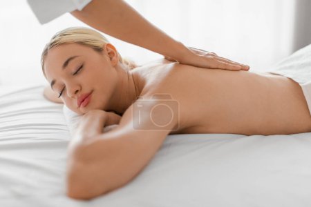 Téléchargez les photos : Une femme blonde détendue s'allonge sur une table de massage dans un spa et reçoit un massage du dos d'un massothérapeute. Le thérapeute utilise une pression ferme et diverses techniques - en image libre de droit