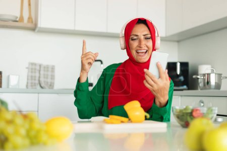 A smiling woman, clad in a vibrant red headscarf and green top, is captured singing and gesturing with her hand as if holding a microphone. She is wearing headphones