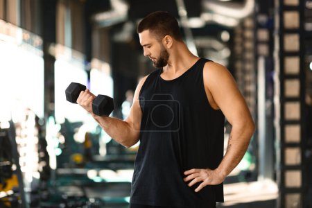 A focused, athletic man with a beard is standing inside a well-equipped gym, wearing a sleeveless shirt as he performs bicep curls with a black dumbbell