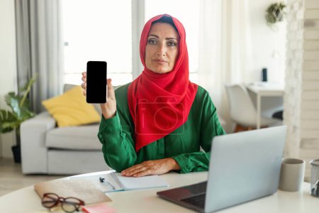 Muslim woman sitting at a table, engrossed in her work on a laptop while also showing her cell phone with blank screen. She appears focused and determined as she multitasks between the two devices.