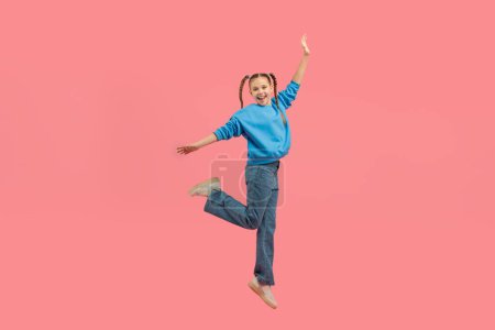 Photo for An energetic teenager girl jumps midair with her hand raised, expressing pure joy, isolated on a pink background - Royalty Free Image