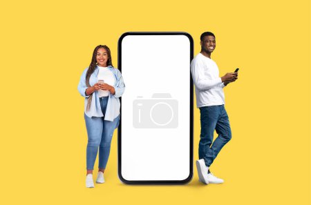 Ein lächelnder junger afroamerikanischer Mann und eine junge Frau flankieren eine große Smartphone-Attrappe auf gelbem Studiohintergrund