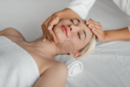 A woman lies on a spa bed while a professional gently massages her face, focusing on pressure points and areas of tension, top view