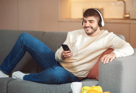 A smiling young man lounges comfortably on a grey couch in a cozy living room setting. He is wearing casual clothing and headphones, and is holding a smartphone, possibly choosing music