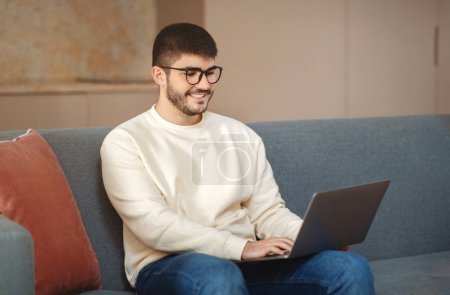 Photo for A man wearing eyeglasses is sitting comfortably on a modern couch, engrossed in his work on a laptop computer, home interior - Royalty Free Image