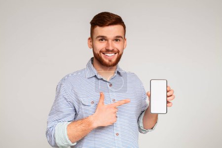 Foto de Sonriente hombre milenario sosteniendo el último teléfono celular delgado con pantalla blanca y apuntando a ella, panorama, espacio libre - Imagen libre de derechos