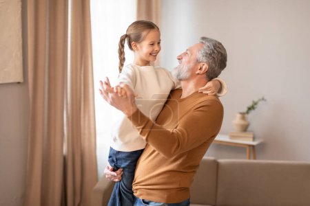 Un hombre mayor está de pie, sosteniendo a una niña sonriente en sus brazos. Ambos parecen felices y conectados, mostrando un momento conmovedor de amor familiar.