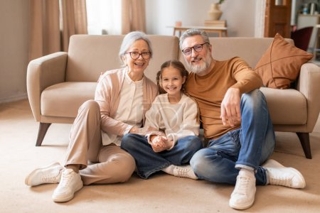 A cheerful elderly couple and their young granddaughter are sitting closely together on the floor of a cozy living room, projecting warmth and family togetherness.