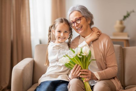 A joyful grandmother with glasses and her young granddaughter, with pigtails, are posing together on a beige sofa. The granddaughter is holding a bouquet of fresh white tulips