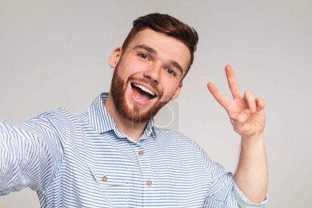 Young millenial man showing V - sign and making selfie on beige background, panorama
