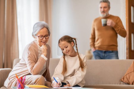 A sweet moment unfolds as a caring grandmother assists her young granddaughter with homework on the living room sofa. The background shows a grandfather looking on warmly with a mug in hand