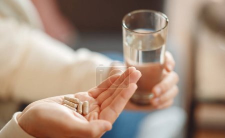 A close-up view illustrates man holding several pills in one hand and clear glass of water in the other, preparing to take medication. The focus on the hands conveys an everyday healthcare routine.
