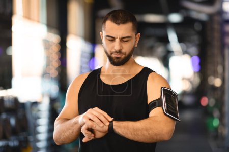 Ein muskulöser Mann in schwarzem Tank-Top und sportlichem Armreif beobachtet sorgfältig seine Fortschritte, indem er auf einen Fitness-Tracker am Handgelenk blickt.