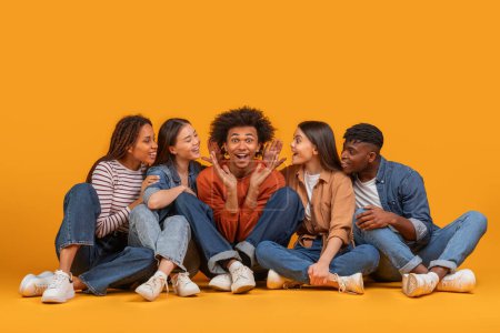 A multiracial group of friends excitedly shares a secret with a young man, indicating trust and closeness in an international, multiethnic setting, isolated on a yellow background