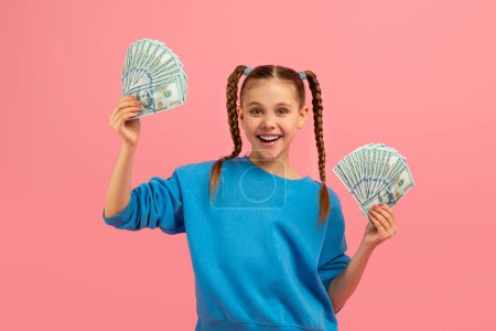 Photo for A young girl with braids smiles while holding up several hundred-dollar bills against a pink background, suggesting wealth or savings - Royalty Free Image