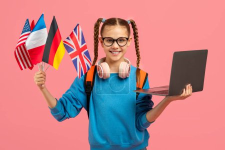 Téléchargez les photos : Une adolescente tech-savvy présentant un ordinateur portable tout en tenant divers drapeaux nationaux, isolé sur une toile de fond rose - en image libre de droit