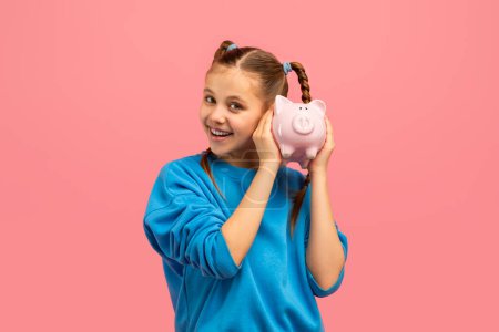 A cheerful teenager girl in a blue sweatshirt holds a piggy bank isolated on a pink background, signifying savings and financial awareness among youngsters
