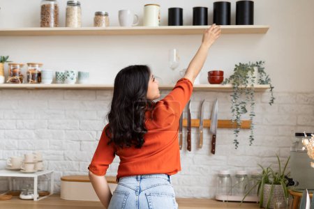In a cozy, well-lit kitchen, a woman in a bright orange shirt is extending her hand to reach items on a shelf