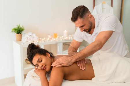 Dans une ambiance spa calme, un thérapeute offre un massage professionnel du dos à une femme noire, illustrant les services thérapeutiques pour les personnes