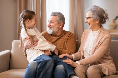 Foto de Un momento reconfortante mientras dos abuelos sonrientes comparten una tierna experiencia con su joven nieta en un acogedor interior de la sala de estar - Imagen libre de derechos