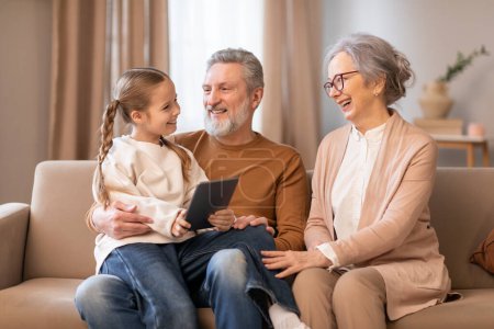 An elderly couple and their young granddaughter are seated together on a beige couch, sharing a moment of joy and connection. The grandfather holds a digital tablet