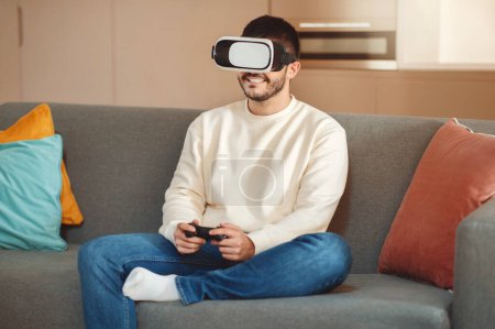 Téléchargez les photos : Un homme est assis sur un canapé, plongé dans une expérience de réalité virtuelle. Il porte un casque VR et semble concentré sur le contenu numérique affiché. - en image libre de droit