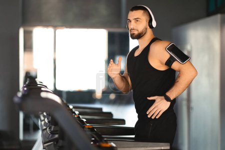 Foto de Un joven enfocado con barba, vistiendo una camiseta negra y auriculares, está junto a una cinta de correr en un gimnasio contemporáneo, listo para comenzar su entrenamiento cardiovascular - Imagen libre de derechos
