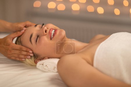 Joyful woman lying down while a professional masseuse gently massages her face, with focus on relaxation and rejuvenation of facial muscles and skin.