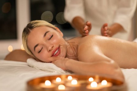 Téléchargez les photos : Joyeux jolie femme se trouve sur une table de massage dans un spa comme un thérapeute massages son dos. Les mains thérapeutes exercent une pression sur les muscles de la femme, apportant soulagement et relaxation - en image libre de droit