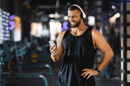 Photo for A man in a sleeveless top and headphones smiling as he selects a playlist on his smartphone in gym. His relaxed posture and joyous expression convey a moment of enjoyment in his fitness routine. - Royalty Free Image