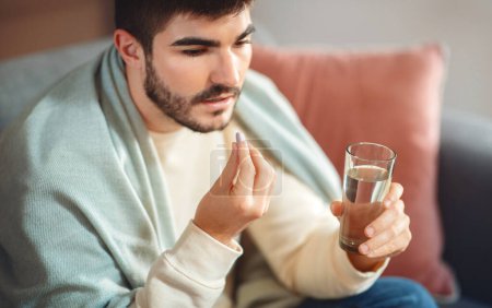 Photo for A focused young man with a short beard, wearing a casual light-colored sweater, is seated while holding a glass of water in one hand and a pill in the other, about to take his medication. - Royalty Free Image