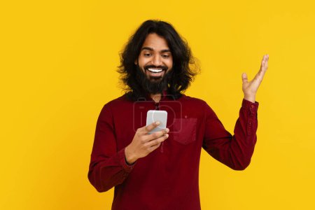 Indian man with long hair and a beard is seen holding a cell phone in his hand. The man appears focused on the screen, possibly texting or browsing, raising hand up