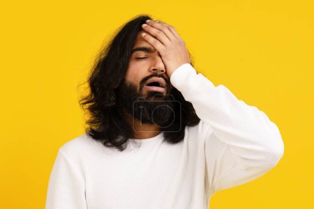 Photo for Indian young man with long dark hair and a beard appears distressed, with his eyes closed and one hand covering his forehead - Royalty Free Image