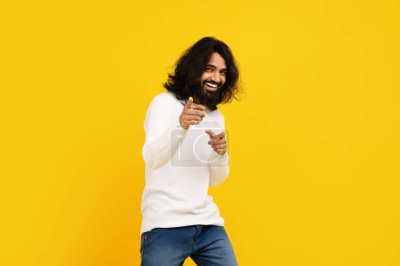 Foto de Hombre indio con el pelo largo y una barba está de pie delante de un fondo amarillo brillante, apuntando a la cámara y sonriendo - Imagen libre de derechos