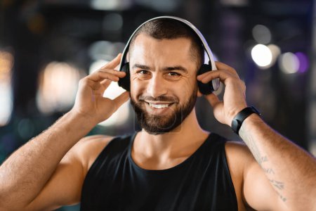 Ein Mann mit Bart trägt Kopfhörer und hört Musik oder Audio. Er wirkt konzentriert oder entspannt, während er das Training im Fitnessstudio genießt, Nahaufnahme