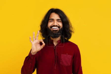 Foto de Hombre indio con barba llena y pelo largo sonríe ampliamente, de pie sobre un fondo amarillo vivo, mostrando gesto OK con su mano derecha, expresando positividad y aprobación. - Imagen libre de derechos