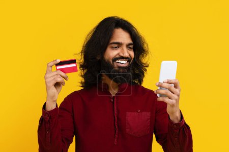 Hombre indio se muestra sosteniendo un teléfono inteligente en una mano y una tarjeta de crédito en la otra. Parece que está haciendo una transacción o compra en línea usando los dos dispositivos.