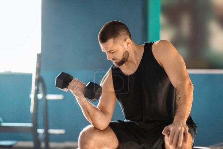 Photo for A man is seated on a bench in gym, gripping a pair of dumbbells in his hands. He appears focused and concentrated on his workout routine, showcasing determination and commitment to fitness. - Royalty Free Image