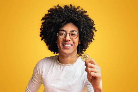 Photo for Cryptocurrency mining. Cheerful millennial black guy holding golden bitcoin, orange studio background - Royalty Free Image