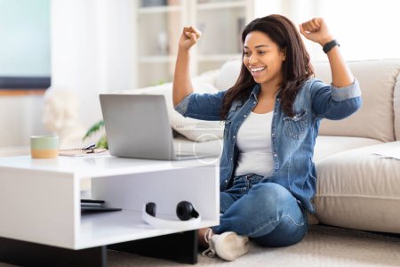 Femme afro-américaine est assise sur le sol avec les jambes croisées, concentrée sur un écran d'ordinateur portable en face d'elle. Elle semble engagée dans le travail ou l'étude, avec une expression heureuse sur son visage.