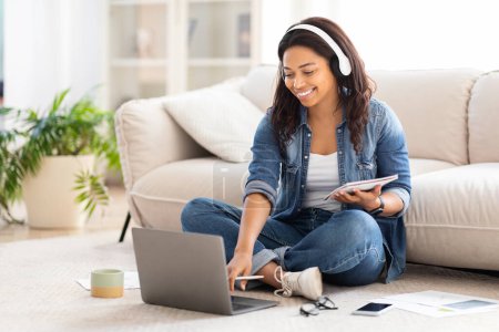 Téléchargez les photos : Femme afro-américaine à l'intérieur, assis sur le sol avec les jambes croisées, concentré sur l'utilisation d'un ordinateur portable devant elle, en utilisant des écouteurs, étudier en ligne de la maison - en image libre de droit