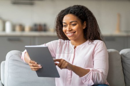 Eine nahbare schwarze Frau hält eine Tablette in der Hand, ihr Lächeln suggeriert Genuss oder Zufriedenheit aufgrund des Inhalts, den sie zu Hause betrachtet.