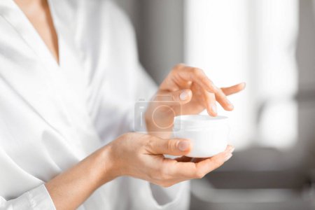 Photo for A detailed close-up captures a woman hands as they gently apply a moisturizing cream from a white jar, focusing on skin care and personal hygiene - Royalty Free Image