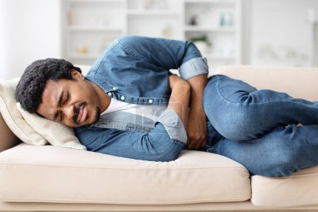 Téléchargez les photos : Jeune homme noir malsain allongé sur le canapé à la maison, touchant son ventre, souffrant de douleurs abdominales - en image libre de droit