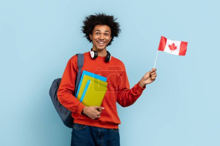 Téléchargez les photos : Un Noir heureux qui fait preuve d'une énergie vibrante et d'une conscience internationale en agitant le drapeau canadien à côté de manuels scolaires brillants, le tout sur un fond clair et bleu isolé - en image libre de droit